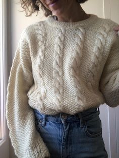 a woman wearing a white sweater and jeans standing in front of a window with her hands on her hips