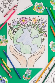 the earth in hands is surrounded by crayons and pencils on a green surface