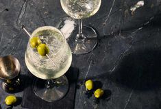 two martini glasses with olives in them on a black counter top next to silverware