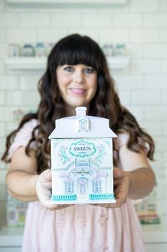 a woman in a pink dress holding up a white house shaped box with the words sweetest on it