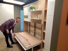 a man standing next to a bed in a room with wooden floors and shelves on the wall