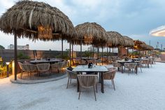 an outdoor dining area with straw umbrellas and tables set up on the sand at dusk