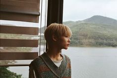 a young man looking out the window at water and hills in the distance with mountains behind him