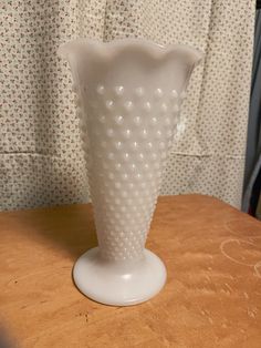 a white glass vase sitting on top of a wooden table next to a wallpapered curtain