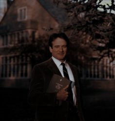 a man wearing a suit and tie holding a book in front of a building with trees