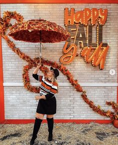 a woman standing under an umbrella in front of a happy fall sign with hay and pumpkins