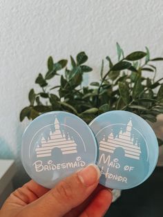 someone holding up two badges that say bridesmaid and maid of the manor in front of a potted plant