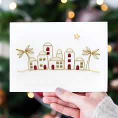 a person holding up a card with houses and palm trees on it in front of a christmas tree