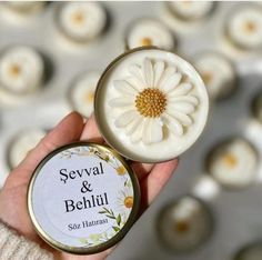 a hand holding a small tin with a flower on it next to some white candles