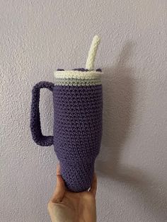 a hand holding a crocheted coffee cup in front of a white wall with a handle