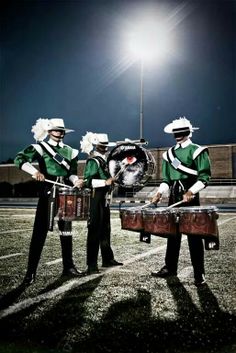 three men in green and white uniforms are playing drums on the football field at night