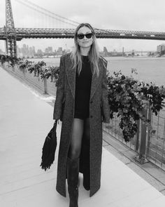 a woman standing on the sidewalk in front of a bridge wearing boots and a coat