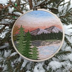 a cross stitch ornament hanging from a tree with snow on the ground and mountains in the background