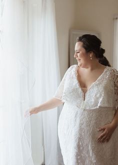 a pregnant woman standing in front of a window with her hand on the sheer curtain