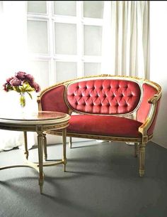 a red couch sitting next to a table with flowers on top of it in front of a window