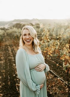 a pregnant woman smiles while standing in a vineyard