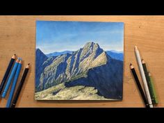 some colored pencils are sitting on a table next to an art work with mountains in the background