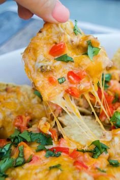 a hand holding a tortilla chip with cheese and tomatoes on it, over a bowl of nachos