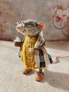 a toy rat dressed in a sweater and glasses is standing on a lace tablecloth