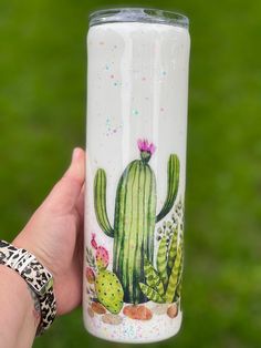 a hand holding a white tumbler with a cactus painting on it's side