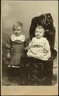 an old photo of two babies sitting on a chair with their heads turned to look like they are looking at the camera