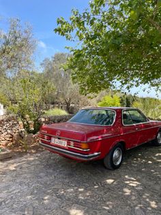 an old red car parked in front of a stone wall with trees and bushes behind it