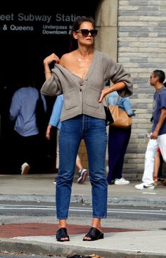 a woman standing on the sidewalk in front of a building wearing jeans and a sweater