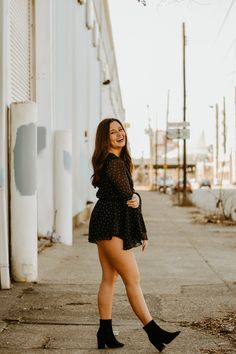 a woman in black dress and boots standing on the side of a road smiling at the camera