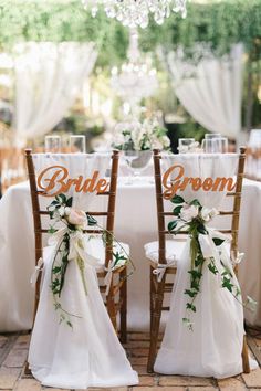 two wooden chairs with white sashes and flowers on them are set up for an outdoor wedding