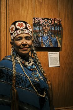 an old native american woman standing in front of a wooden wall with pictures on it