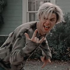 a man with blonde hair making the peace sign in front of a house and bushes