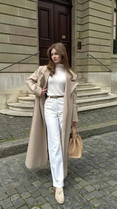 a woman in white pants and a coat is standing on a brick sidewalk with her hands behind her back