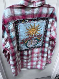 a pink and white plaid shirt with an embroidered sun on the chest, in front of a door