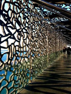 people are walking down a long walkway next to the water and an intricate metal wall