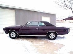 an old purple car parked in front of a garage with snow on the ground next to it