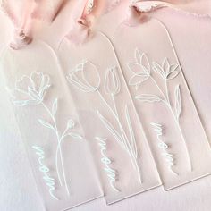 three tags with flowers drawn on them sitting next to a pink ribbon and some fabric