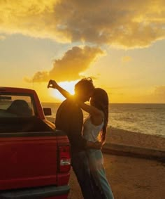 a man and woman standing next to a red truck on the beach at sun set