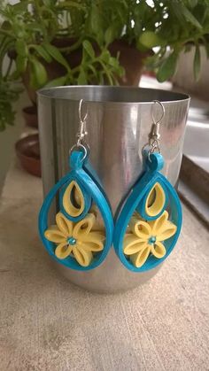 two blue and yellow earrings sitting on top of a metal container next to a potted plant