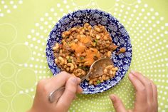 two hands holding a spoon over a blue and white bowl filled with food on top of a green tablecloth