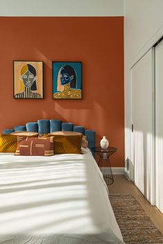 a bedroom with orange walls and white bedding, two paintings on the wall above it