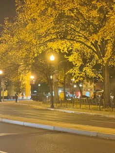 an empty street at night with yellow leaves on trees and people walking down the sidewalk