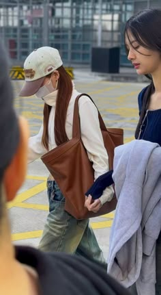 two young women are walking together in an airport, one carrying a brown bag and the other wearing a white hat