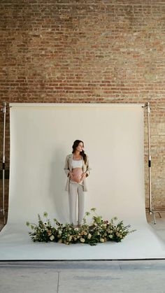 a woman standing in front of a white backdrop with flowers and greenery on it