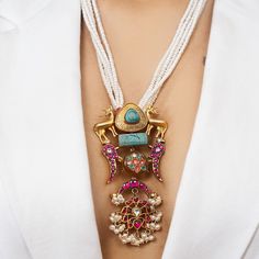 This Jadau Necklace is crafted to make a bold statement, showcasing a dramatic pendant with a diverse range of motifs and stones. The striking pendant features a plain metal deer adorned on both sides with a beautiful turquoise stone in the center. Intricate parrot, crescent-shaped motifs, and delicate florals enhance the pendant, all embellished with premium quality polki and red stones in the traditional Jadau craft style. The pendant is suspended from a series of pearl strands, and the bottom Craft Necklace, Jadau Necklace, Jaipur Jewelry, Jadau Jewellery, Antique Necklaces Design, Antique Necklaces, Red Stones, Pearl Necklace Designs, Silver Jewellery Indian