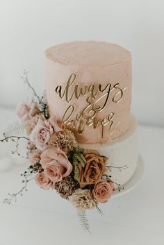 a wedding cake with pink frosting and gold lettering on top, surrounded by flowers