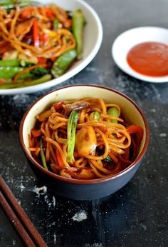 two bowls filled with noodles and vegetables next to chopsticks on a black table