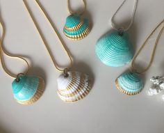 four different seashells are hanging from gold chains on a white surface, one is blue and the other is green