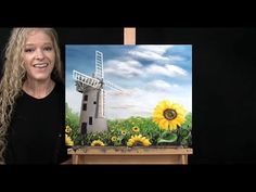 a woman standing next to a painting of a windmill and sunflowers