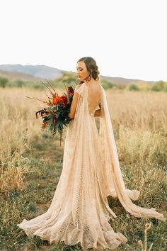 a woman standing in a field wearing a long dress and holding a flower bouquet with her back to the camera