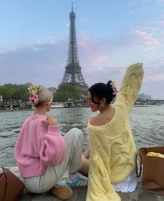 two women sitting next to each other near the water in front of the eiffel tower